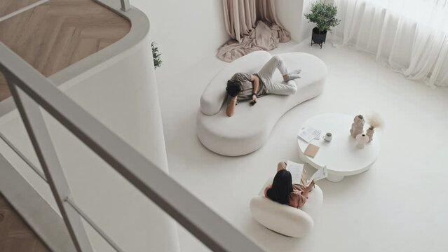 High angle of young man lying on couch, using smartphone, dark-haired woman sitting in armchair, working with documents in living room with modern interior design at daytime