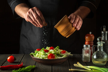 The chef sprinkles salted fresh vegetable salad in a plate on a wooden table. Cooking healthy food in the kitchen in a restaurant.