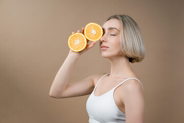Slim woman with beautiful face complexion holding oranges halves in hands. Model in white lingerie posing on beige background, advertising healthy lifestyle and fitness concept.