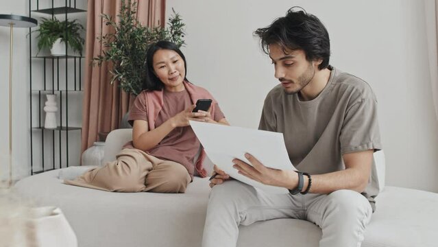 Young Middle Eastern man holding papers, talking to Asian woman who using smartphone, couple sitting on couch in living room with modern interior design at daytime