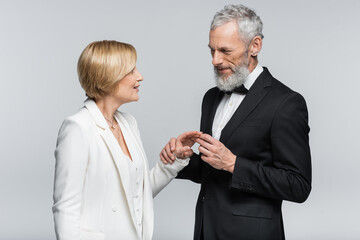 Middle aged groom wearing wedding ring on smiling bride isolated on grey.
