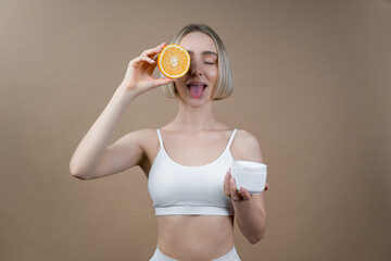 Slim woman with beautiful face complexion holding an orange half and cream container. Model in white lingerie posing on beige background, advertising beauty and fitness concept.