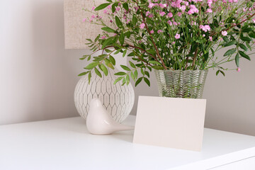 Blank paper card, vase of spring flowers, home decor on white table. Happy Mothers Day greeting card template in scandinavian style cozy home living room interior