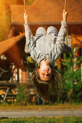Young beautiful girl hanging upside down and having fun on a swing outdoor. Happy childhood, summer vacation, holiday and rest concept. Vertical image.