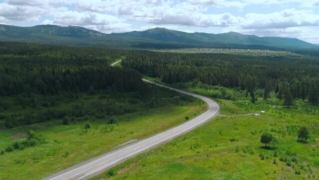  A long road with cars. The view from the drone. Scene. A bright summer landscape with seedy mountains and forest in the background next to a road with cars in summer against a bright sky.