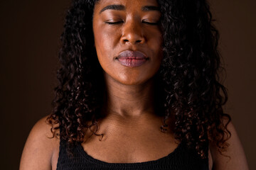 Mid waist portrait of african american woman with eyes closed having meditation. Vertical front view of african woman breathing with relax expression in brown background. People and emotions concept.