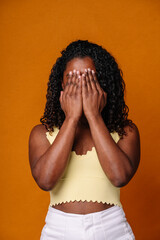 Mid waist portrait of african american woman covering her eyes in orange background. Vertical front view of african woman covering her face with depression. People and emotions concept.