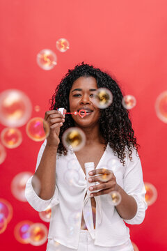 Mid waist portrait of cheerful woman blowing soap bubbles in red background. Vertical front view of african american woman playing funny with soap bubbles. People background concept.