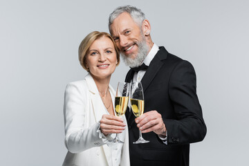 Smiling mature groom looking at camera near bride with champagne isolated on grey.