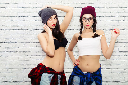  Stylish sexy hipster twin girls in baseball caps, bare midriff crop top, short jeans and sunglasses in studio. Two trendy urban teenage girls spend time making faces together.