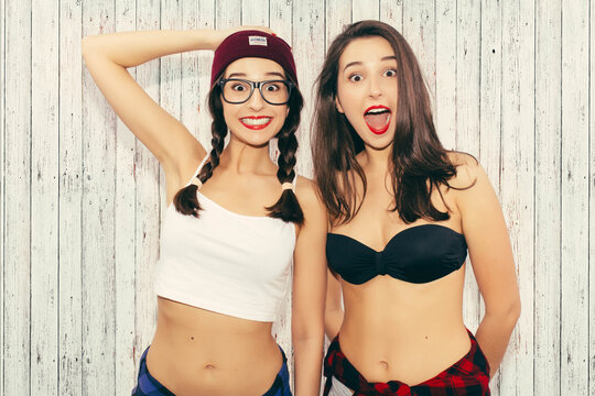 Stylish hipster twins girls in hats, wearing short top with naked belly, jeans short and sunglasses in the studio. Two fashion urban teenagers women spend time together having fun make funny faces.