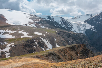 Vivid landscape near precipice edge with sharp rocks with view to large snowy mountains and glacier under cloudy sky. Picturesque view from stone hill to high mountain range with shadows from clouds.