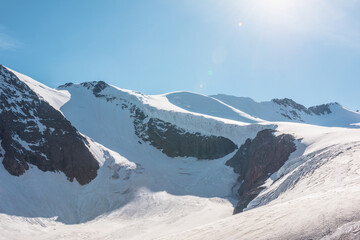 Minimalist mountain landscape with large glacier in sunlight. Simple snowy minimalism with glacial tongue. Minimal alpine view to snow mountain tops at very high altitude under blue sky in sunny day.