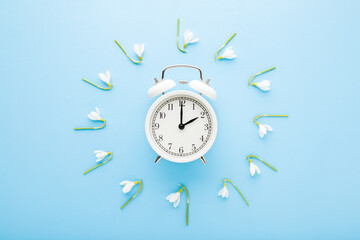 Fresh beautiful snowdrops  around white alarm clock on light blue table background. Pastel color. First messengers of spring. Time change concept. Closeup. Top down view.