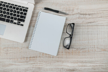 Top view photo of laptop, glasses, notepad and pen on light wooden table background