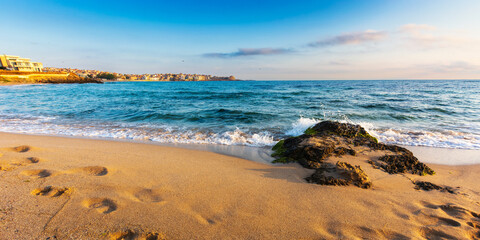 calm morning scenery at the beach. wonderful summer vacation background. calm waves washes the shore. blue sky with some clouds glowing in morning light. steps on the sand. seaweed on the stone