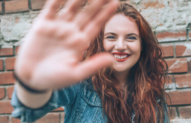 Fashion portrait of sincerely smiling red curled long hair caucasian teen girl with applied red lipstick lips with blue eyes with a red brick wall background doing a "No photo" gesture.