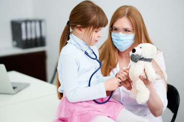 Caucasian female doctor wear face mask show little girl examining bear toy by stethoscope indoor hospital