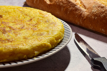 Potato omelette, typical of Spain, on a porcelain plate, stone background, along with a knife and bread.