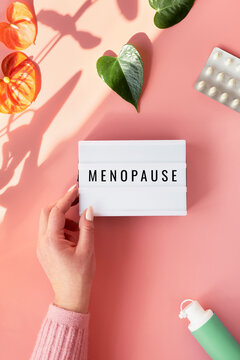 Text Menopause on light box in hand. HRT Replacement hormone therapy, HRT concept. Pink background with exotic leaves, flowers, pills, estrogene gel. Sunlight, long shadows.
