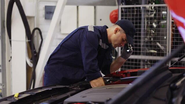 Car service worker examinating engine under hood with flashlighter in hand, side view
