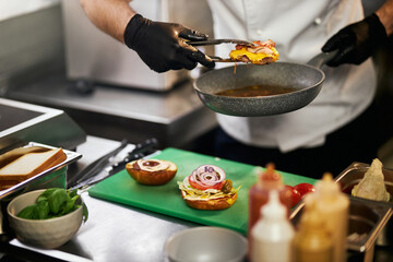 Crop view of male hands in gloves holding juicy grilled putty with cheese and bacon, using tongs...