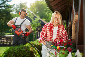 Strong caucasian man in uniform using electric trimmer for pruning bushes at garden while charming woman cutting dry leaves on flower in pots. Seasonal work outdoors. 