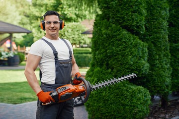Smiling caucasian man in overalls, safety glasses and gloves holding electric hedge trimmer while...