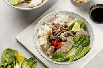 Pho Bo vietnamese soup with beef, rice noodles, pak choi and mushrooms in a grey bowl on marble board, peanuts, soy sauce, chilli