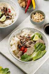 Pho Bo vietnamese soup with beef, rice noodles, pak choi and mushrooms in a grey bowl on marble board, peanuts, soy sauce, chilli