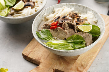 Pho Bo vietnamese soup with beef, rice noodles, pak choi and mushrooms in a grey bowl on wooden board, peanuts, soy sauce, chilli