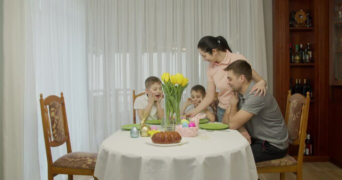 Cheerful mother brings a basket of painted eggs and puts it on the table while serving the holiday dinner for a large family