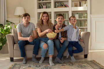 Happy family, a woman and three boys, are sitting on the sofa in the living room with a bowl of popcorn, watching TV