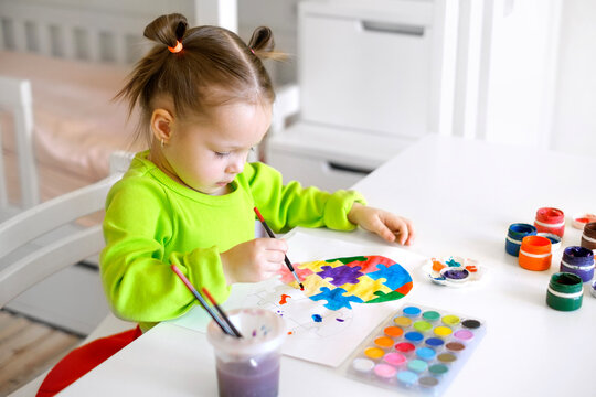 Kid is concentrating on coloring the puzzle pieces inside the heart with different colors. Girl diligently draws a drawing as a present to friends with autism syndrome. Gouache and watercolor paints