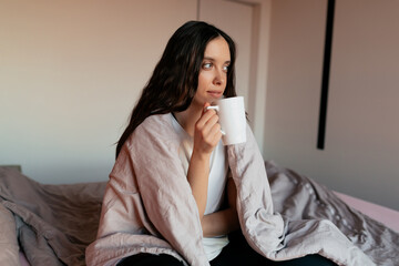 Pretty happy stylish girl with wavy dark hair is sitting in the bed with morning coffee. Lovely...