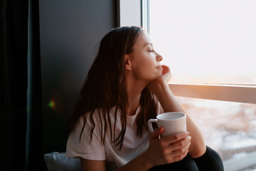 Close up portrait of romantic lovely woman with dark hair with wavy hair is closed eyes and smiling while drinking morning coffee and sitting near the window in morning sunshine