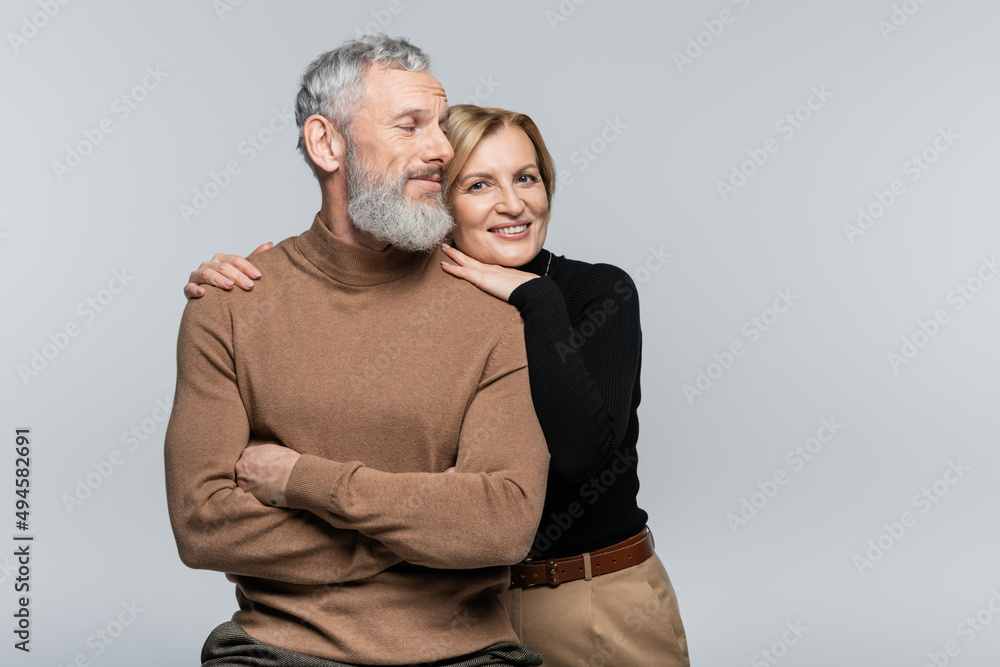 Wall mural smiling mature woman in turtleneck hugging husband and looking at camera isolated on grey.