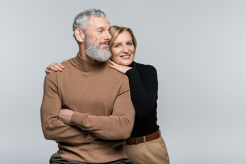 Smiling mature woman in turtleneck hugging husband and looking at camera isolated on grey.