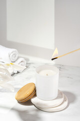 An aromatic soy wax candle is set on fire with match. An ecological and vegan candle, bath accessories and a wooden cap on a marble table. Aromatherapy, scent, relax. Still life composition concept