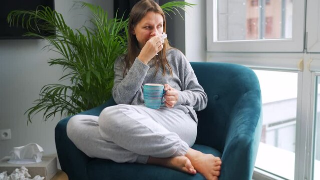 Unhealthy woman sits in a chair, drinks hot tea or a cold medicine and sneezes or blows her nose into a napkin because she has a cold, flu, coronavirus. It is snowing outside