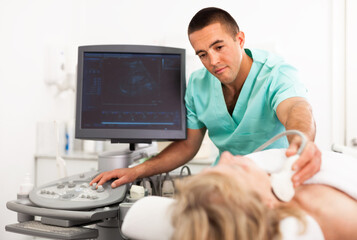 Young positive smiling man sonographer using ultrasonography machine checking female patient in hospital diagnostic room