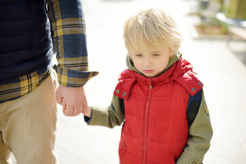Man with sad little boy are walking hand in hand along city street. Frustrated child does not want...