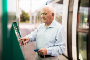 Senior man using ATM machine with credit card while waiting for tram on station.