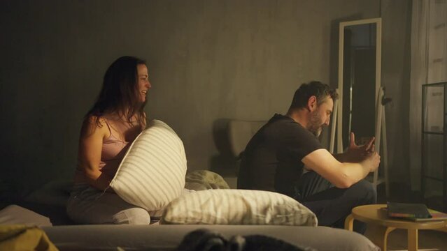Jealous betrayed woman beating her husband with pillow in bed, relationship problems concept.