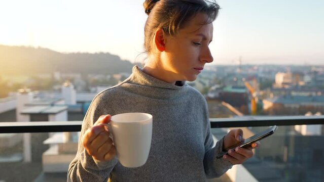 Woman starts her day with a cup of tea or coffee and checking emails in her smartphone on the balcony at dawn, slow motion.