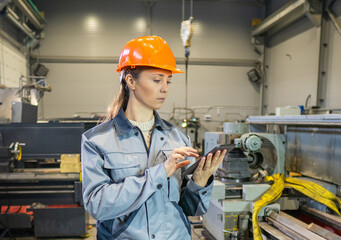 woman in overalls and hard hat holds a tablet and looks at it. factory, production. portrait