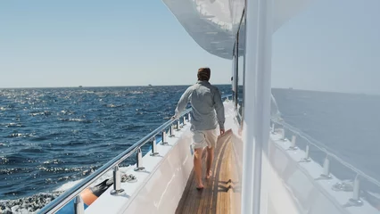 Foto auf Acrylglas Man walks on moving yacht deck on sunny summer day. Caucasian man in light clothes, barefoot on white yacht sailing by blue sea, clear sky above horizon. Luxury lifestyle vacation private sea cruise © krovsmolokom13