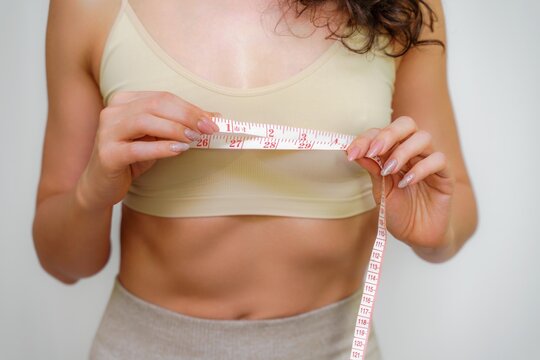 Cropped view of slim woman measuring breasts with tape measure at home, close up. Unrecognizable European woman checks the result of a weight loss diet or liposuction indoors. Healthy lifestyle.