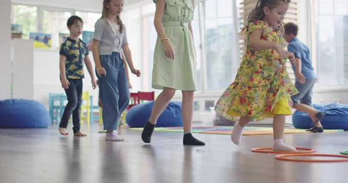 Small nursery school children with female teacher on floor indoors in classroom, doing exercise. Jumping over hula hoop circles track on the floor.