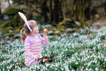 Little girl with Easter bunny ears making egg hunt in spring forest on sunny day, outdoors. Cute...